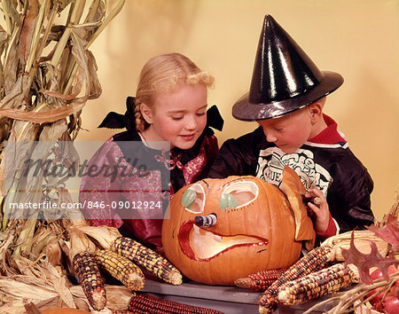 1960s TWO KIDS BLOND BROTHER AND SISTER WEARING HALLOWEEN COSTUMES LOOKING INTO CARVED JACK-O-LANTERN PUMPKIN