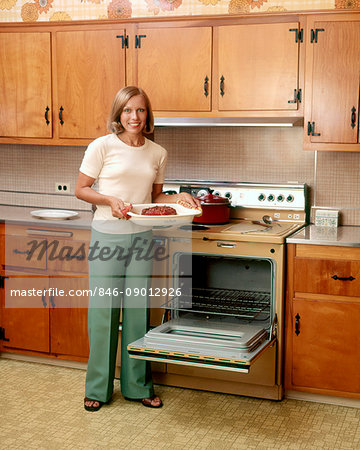 1970s SMILING BLOND WOMAN HOUSEWIFE LOOKING AT CAMERA HOLDING BAKED HAM STANDING BY STOVE OPEN OVEN DOOR
