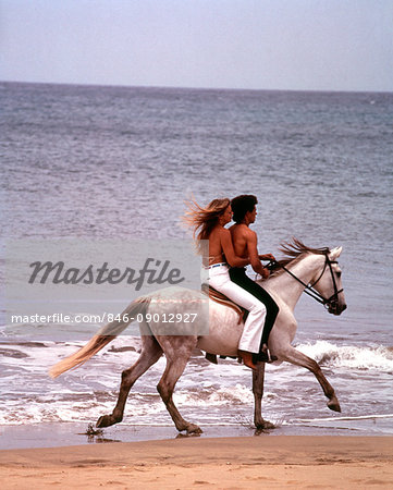 1970s MAN WOMAN COUPLE RIDING TOGETHER ON SINGLE WHITE GRAY HORSE ON BEACH
