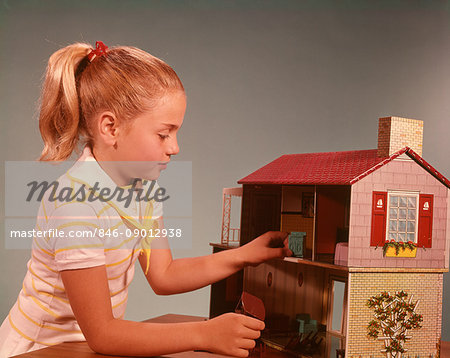 1960s GIRL WITH BLONDE PONYTAIL PLAYING WITH TOY DOLLHOUSE