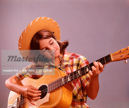 1960s TEENAGER GIRL WEARING COWBOY HAT AND PLAID SHIRT PLAYING GUITAR