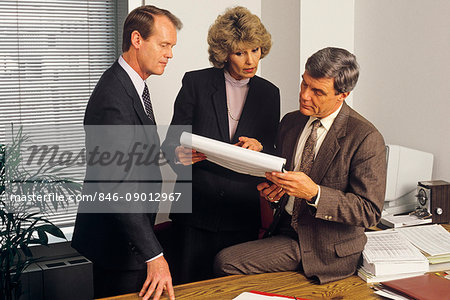 1980s 3 EXECUTIVES HOLDING MEETING IN OFFICE REVIEWING PAPER WORK SPREAD SHEET