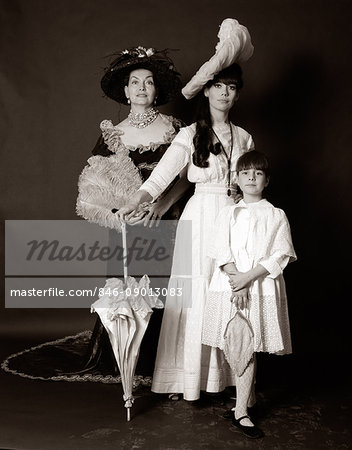 1960s THREE GENERATIONS WOMEN GRANDMOTHER MOTHER DAUGHTER POSED STANDING LOOKING AT CAMERA WEARING 1890s FASHION DRESSES