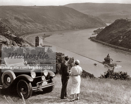 1930s COUPLE WITH TOURING CAR OVERLOOKING RHINE RIVER BURG GUTENFELS AND CASTLE PFALZGRAFENSTEIN ON FALKENAU ISLAND GERMANY