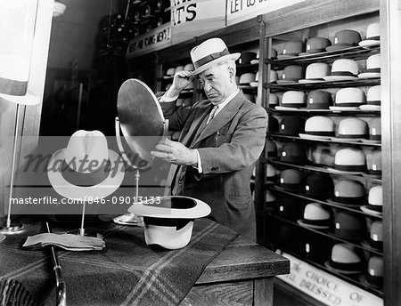 1940s SENIOR MAN TRYING ON HATS LOOKING IN MIRROR IN HAT STORE