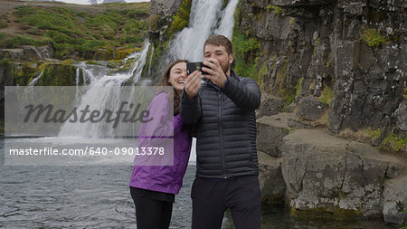 Boyfriend and girlfriend posing and taking smartphone selfie near waterfall over remote rock cliffs