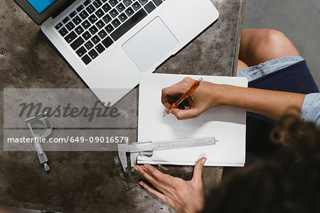 Overhead view of female jeweller's drawing in notepad at workbench