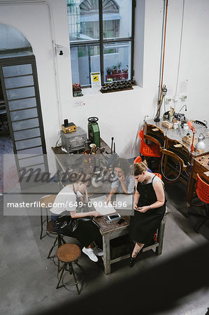 High angle view of three female jewellers looking at sketchpad at workbench meeting