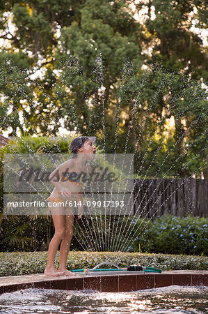 Young girl trying to drink water from sprinkler beside outdoor swimming pool