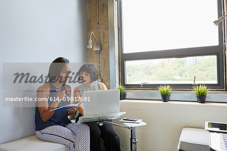 Woman working on laptop, on laptop stand, colleague sitting beside her writing in notebook