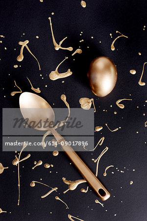 Overhead view of gold painted easter egg and spoon with  splatters on black background