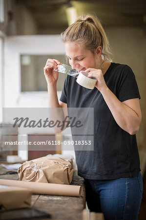 Woman standing in a workshop using her teeth on a piece of sticky tape off a roll, packets and brown packaging paper on a table.