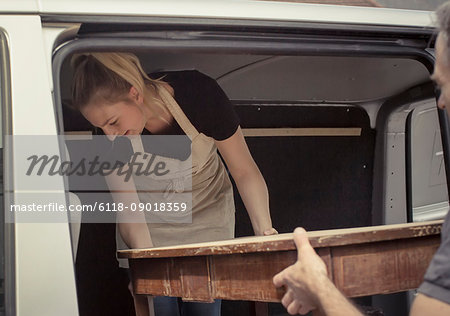 Two people, a man and a young woman lifting an antique wooden table into the back of a van.