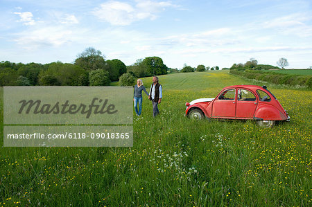 Man and woman walking hand in hand across a meadow, vintage red car parked close by.