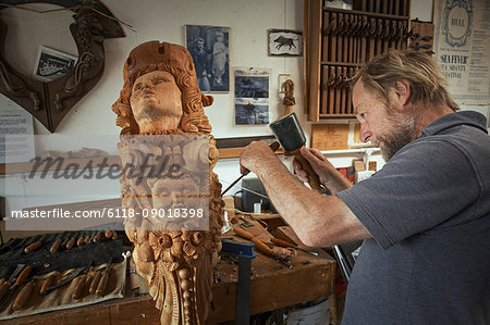 A craftsman, carver working on a carving with a mallet and small chisel marking the wood and adding texture and shape.  A wooden female ship's figurehead, work in progress. Hand tools on the bench.