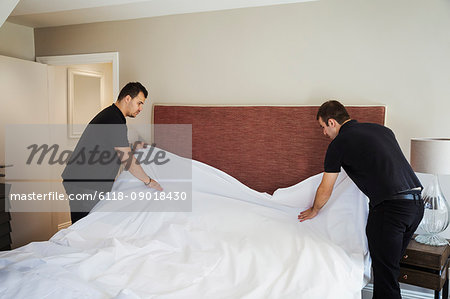 Two men standing in hotel room, making bed.