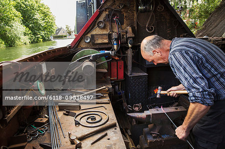 Blacksmith working in a small space on his narrowboat, a barge on river, bending over the anvil and shaping hot metal.