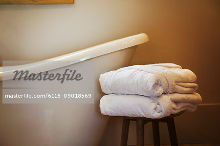 An old fashioned slipper shape bathtub, bath with raised end and wall mounted taps in a bathroom. Two folded guest bathrobes on a stool.