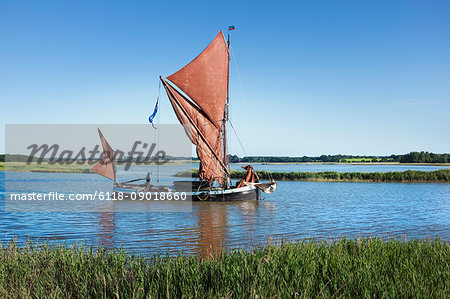 Traditional sailing boat with red sails and a gaff rig, a sailing smack on the water on a reed bed or river estuary.