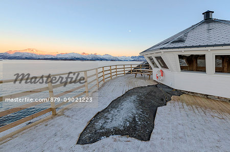 Wooden walkway and terrace on the cold sea surrounded by snowy peaks at sunset Hamn i Senja Bergsfjordens Tromsø Norway Europe