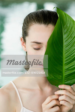 Young woman holding large leaf over half of her face, portrait