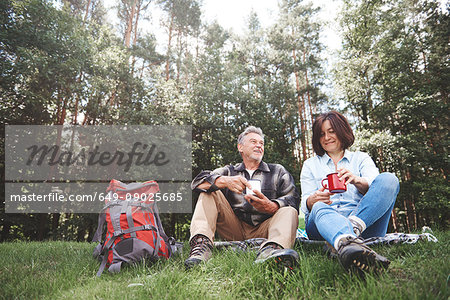 Mature couple relaxing on grass, holding tin cups, rucksack beside them