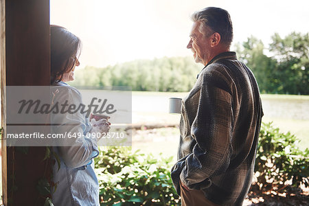 Mature couple standing on veranda, holding tin cups, smiling