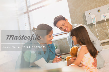 Smiling female nurse using stethoscope on girl patient in hospital room