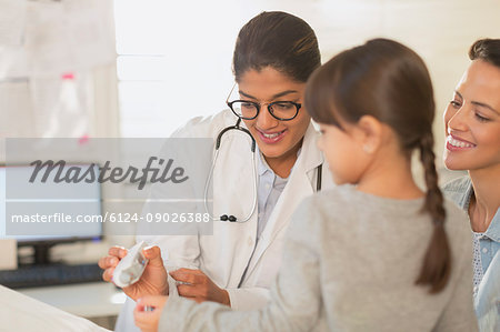 Female pediatrician showing digital thermometer to girl patient and mother in examination room