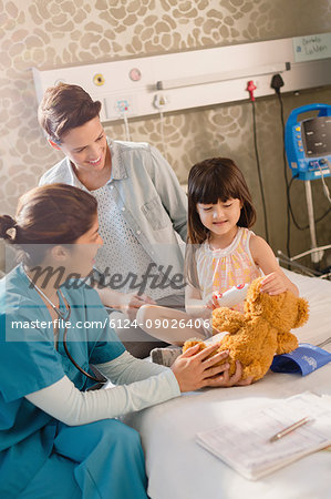 Female nurse and girl patient using digital thermometer on teddy bear in hospital room