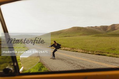 Car window view of young male skateboarder skateboarding along rural road, Exeter, California, USA