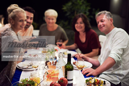 Group of people sitting at table, enjoying meal, young woman taking selfie of group using smartphone