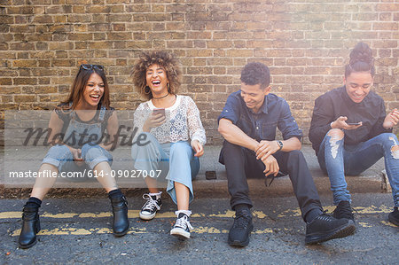 Four friends sitting in street, laughing, young woman holding smartphone