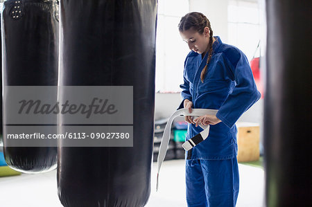 Young woman tying judo belt at punching bags in gym