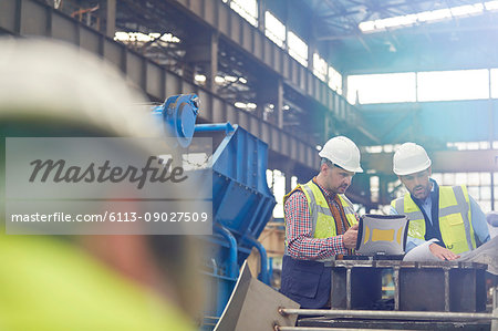 Male foreman and engineer working at laptop, discussing blueprints in factory