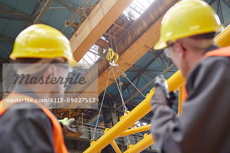 Male worker using walkie-talkie to guide hydraulic crane lowering equipment in factory