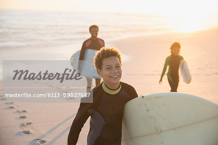 Portrait smiling pre-adolescent boy in wet suit carrying surfboard on summer sunset beach with family