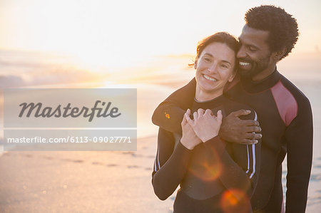 Smiling, affectionate multi-ethnic couple in wet suits on sunny summer sunset beach