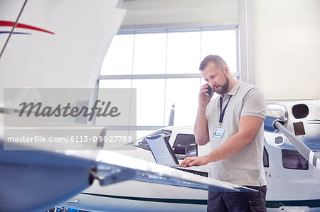 Male airplane mechanic talking on cell phone and working at laptop in hangar