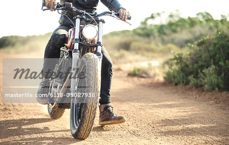 Low section view of man riding cafe racer motorcycle on a dusty dirt road.