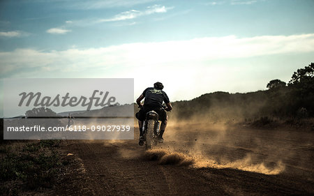 Rear view of man riding cafe racer motorcycle on a dusty dirt road.