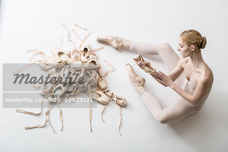 Young ballerina in white tights is sitting on the floor and holding a ballet slip in her hands. Before her a pile of pointe shoes in different condition. Studio shooting, horizontal top shot.