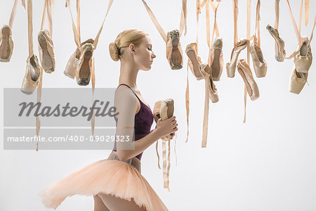 Sensual ballerina in a violet top and a peach tutu stands sideways on the light background in the studio. Around her there are many hanging beige pointe shoes. She holds one pair in the hands.
