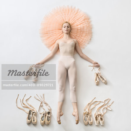 Blonde ballerina in light dance wear with closed eyes lies with stretched legs on the peach tutu on the floor and holds pointe shoes in the studio. On the sides of her legs there are ballet shoes.