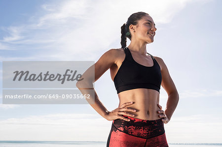 Young female runner on beach with hands on hips against blue sky