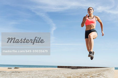 Young woman running along sea wall, mid air