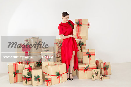 Young woman in red dress by stacked christmas gifts holding christmas parcel
