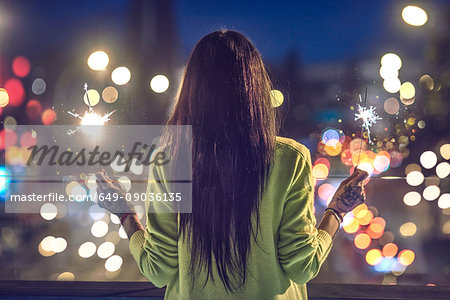 Young woman outdoors at night, holding lit sparklers, tattoos on hands, rear view