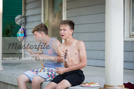 Two teenage boys sitting on porch, eating and smoking cigar