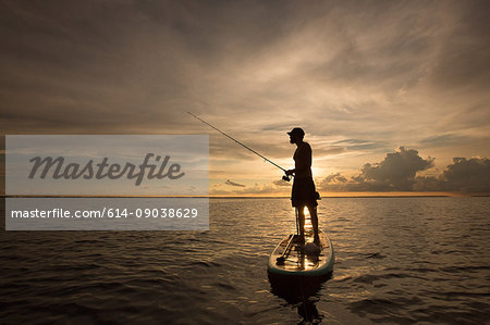 Man standing on paddle board, on water, at sunset, holding fishing rod
